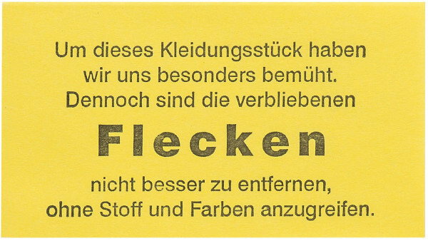 Detailansicht des Fleckenzettels gelb , Beschriftung : Um dieses Kleidungsstück haben wir uns besonders bemüht, dennoch sind die verbliebenen Flecken nicht besser zu entfernen ohne Stoff und Farben anzugreifen